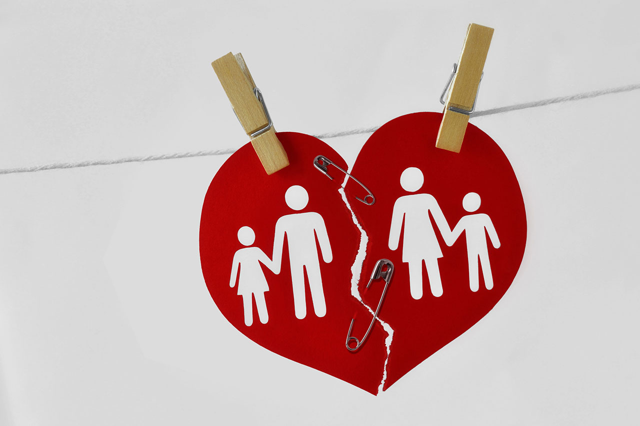 Torn red heart paper hanging on a line with wooden pegs, divided into two halves showing separated family figures of a father and daughter on one side and a mother and son on the other, secured with safety pins, symbolising divorce.